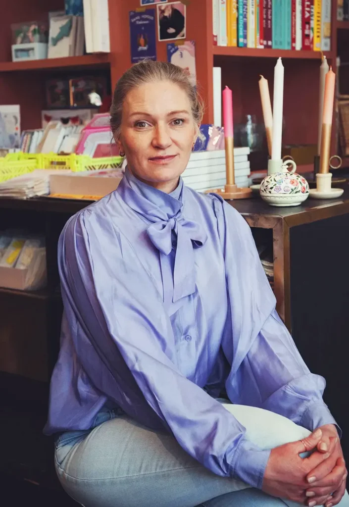 Silke sitzt mit überschlagenen Beinen vor einem Regal mit Büchern Keramik und Kerzen und blickt in die Kamera