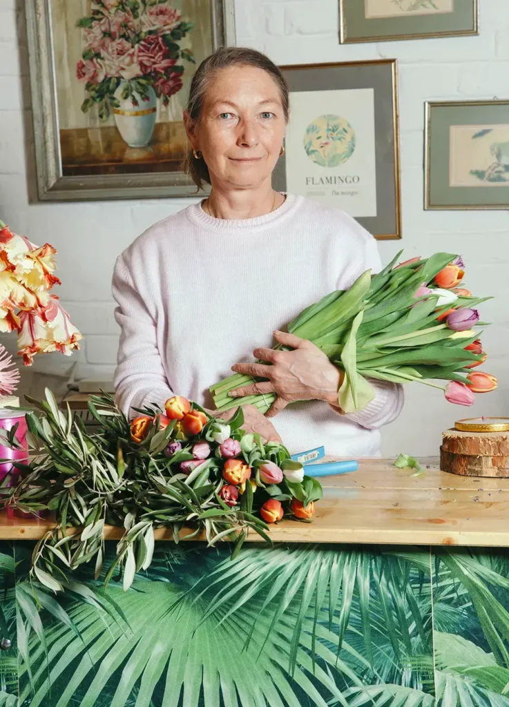 Iris steht mit dem Arm voller Tulpen hinter einer Theke aus Holz, auf der weitere Blumen und Arbeitsgeräte liegen. Im Hintergrund eine Wand mit gerahmten floralen Prints.