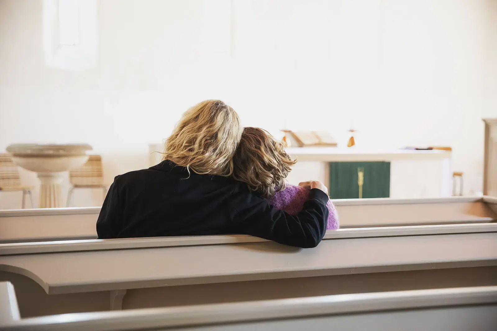 Rückansicht von zwei Personen, die bei einer Bestattung in einem hellen Raum auf einer Kirchenbank sitzen. Eine hat den Arm um die andere gelegt. Im Hintergrund sind Altar, Taufbecken und Kreuz sichtbar.