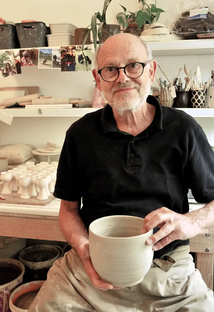 Der Töpfer Hansjörg Schwarz in seinem Atelier. Er hat ein gedrehtes Tongefäß in der Hand und blickt in die Kamera. Im Hintergrund regale mit Töpferarbeiten und Utensilien.