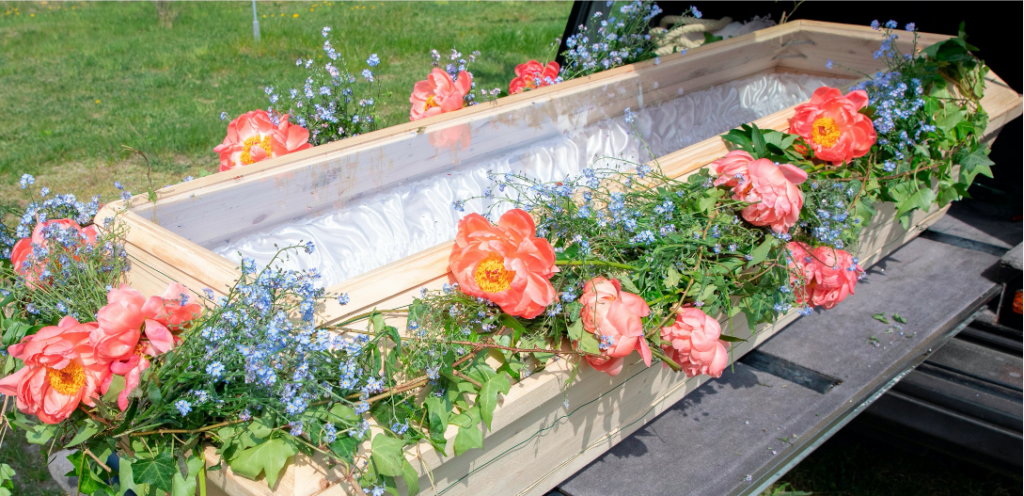 Ein Sarg mit einem durchsichtigen Deckel, der rundherum mit Blumen geschmückt ist, steht auf der ausziehbaren Lade eines Leichenwagens. Man kann durch den Deckel die weiße Innenauskleidung des Sarges sehen.