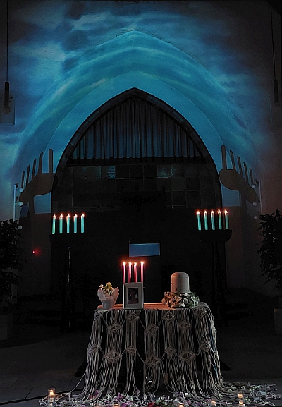 Das Innere einer Kapelle, auf einem Podest ist eine Urne aufgestellt mit Kerzen und Blumen. Der Raum ist dunkel, an die Wand ist eine schillernde Wasseroberfläche projiziert.