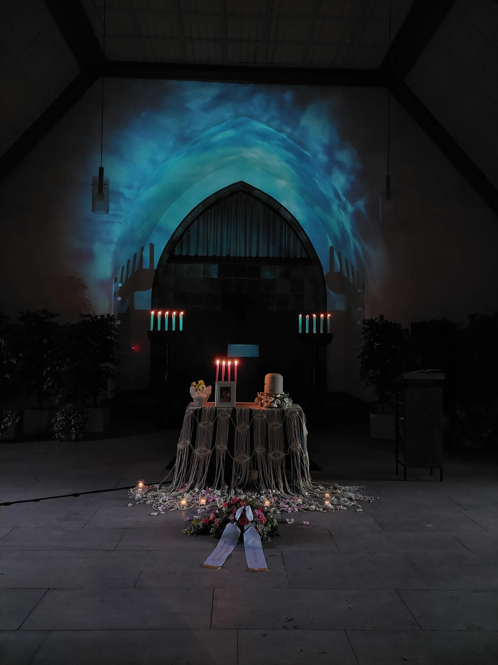 Eine mit blauem Licht beleuchtete Friedhofskapelle. Im Vordergrund steht ein Tisch mit einer Urne und ein Bild der Verstorbenen. Der Tisch ist mit einem weißen Macrame bedeckt. Das ist eine Trauerfeier von Atelier Magnolia Bestattungen in Berlin