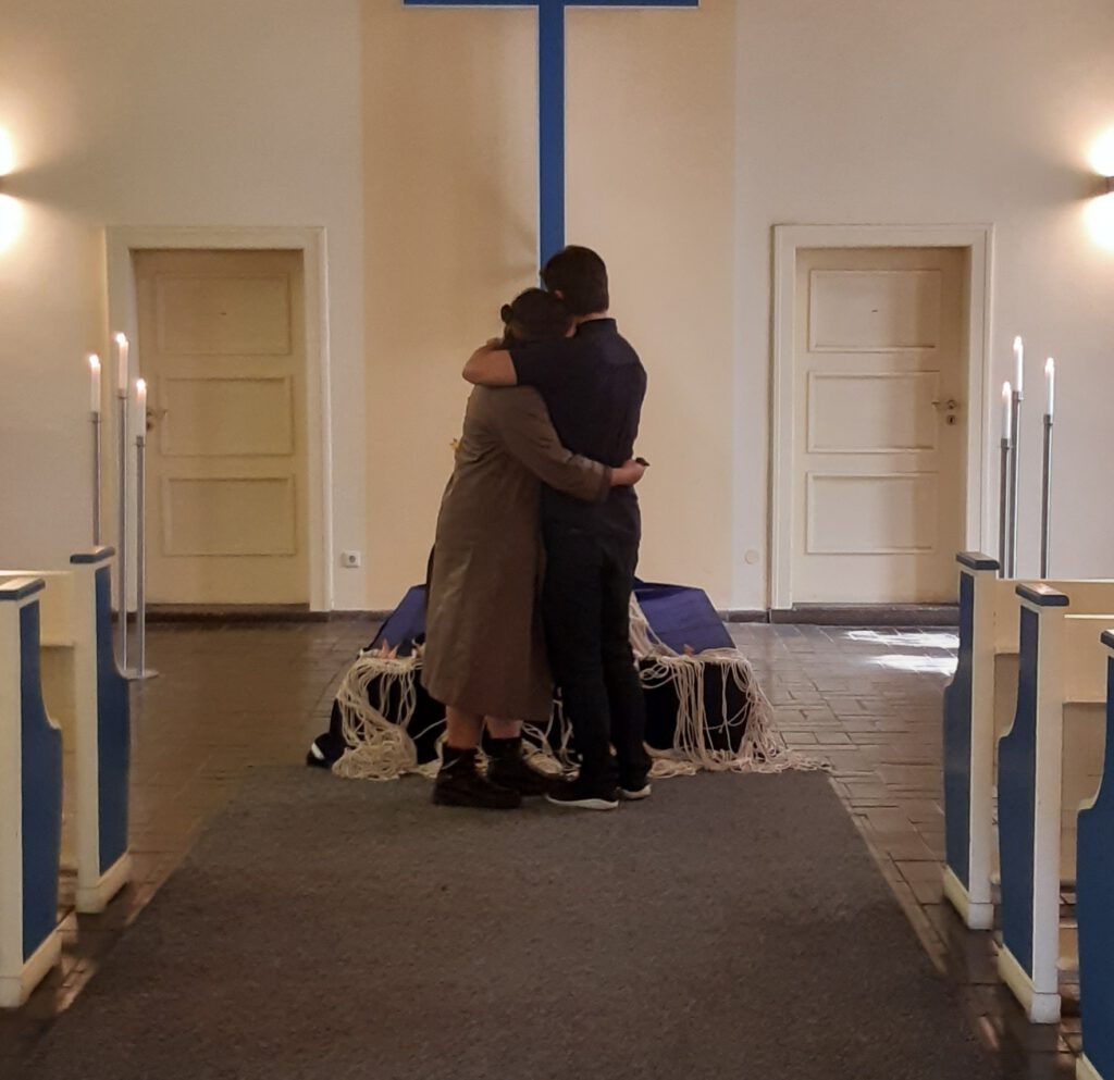 Zwei Menschen von hinten zu sehen, in einer leeren Kapelle, umarmen sich und unterstützen sich damit in ihrer Trauer