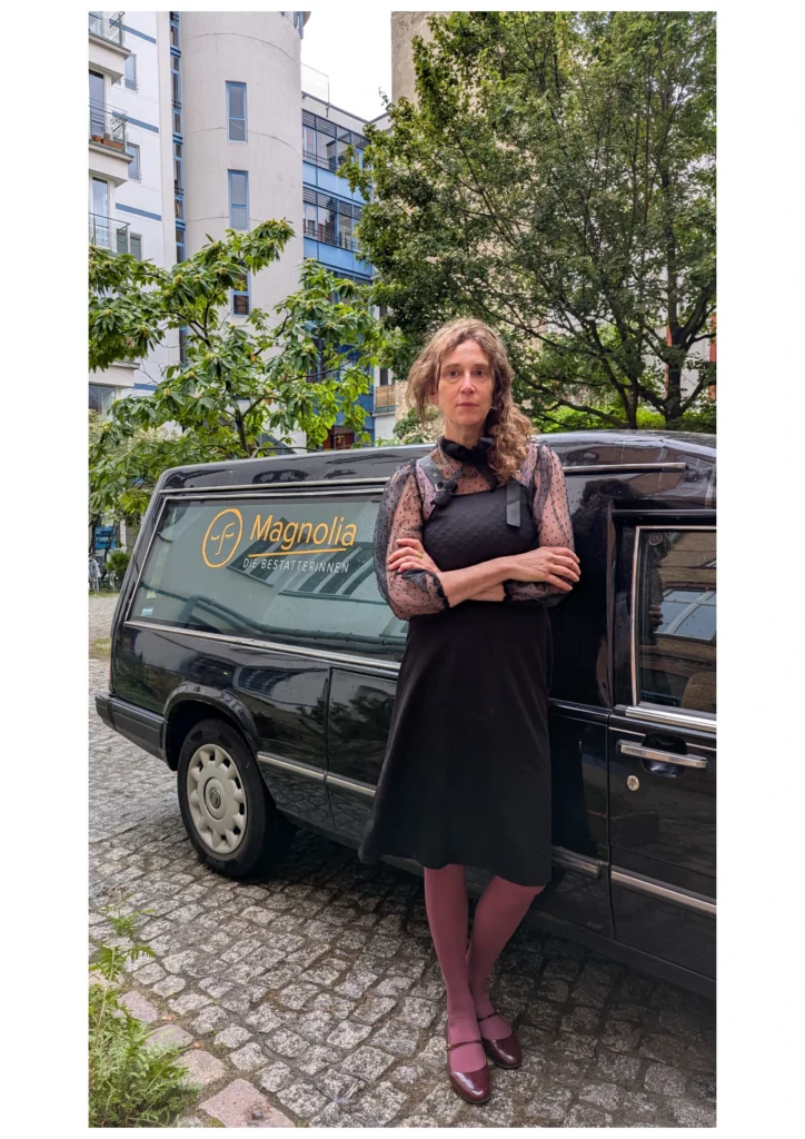 Helena von Atelier Magnolia Bestattungen lehnt an den schönen Leichenwagen und schaut ernst in die Kamera