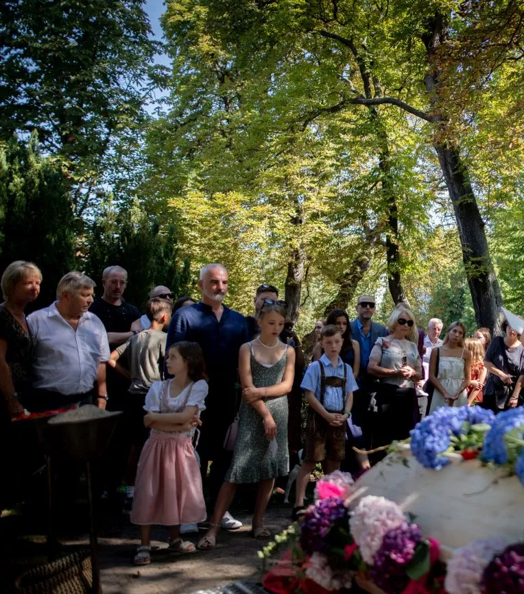 Eine Beisetzung von Magnolia Bestattungen. Eine bunt gekleidete Trauergesellschaft auf einem Friedhof im Sommer. Im Vordergrund des Bildes sieht man einen Teil des Sarges. Es ist ein heller Holzsarg, der üppig mit Hortensien geschmückt ist. Die Trauergesellschaft steht unter grünen Bäumen, ganz vorne stehen drei Kinder.