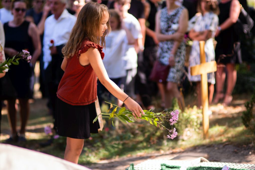 E ein kleines Mädchen steht auf einem Friedhof und wirft Blumen in ein Grab. Hinter ihr steht die Trauergesellschaft.