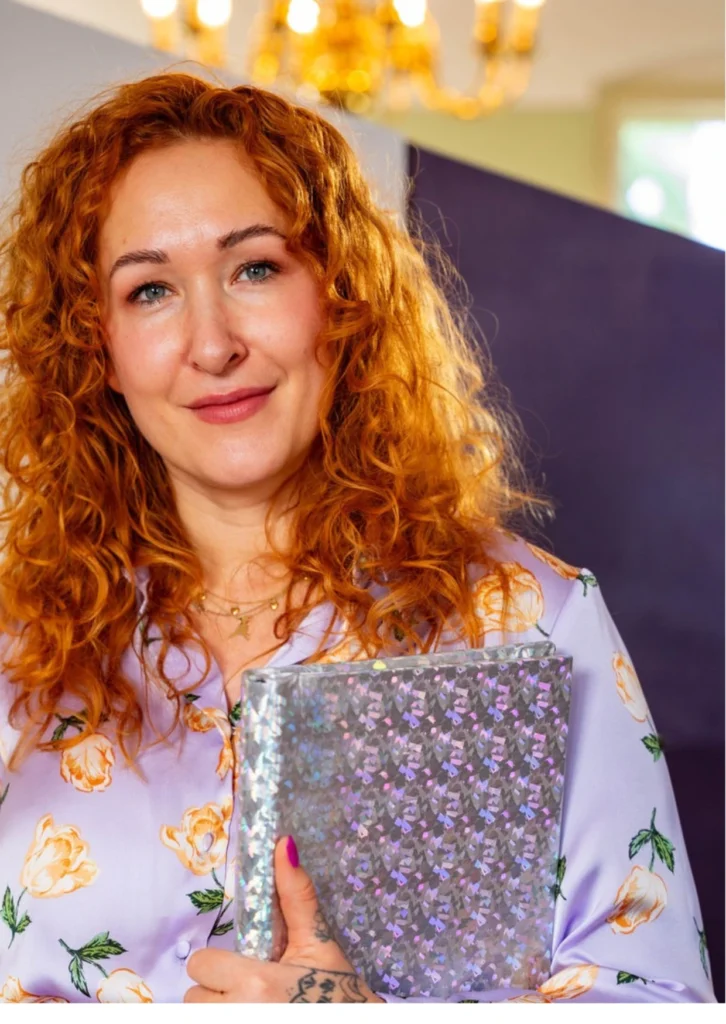 Eine lächelnde Frau mit roten Locken hält ein Buch in der Hand und schaut sehr empathisch. Die Trauerrednerin Liz von Atelier Magnolia Bestattungen