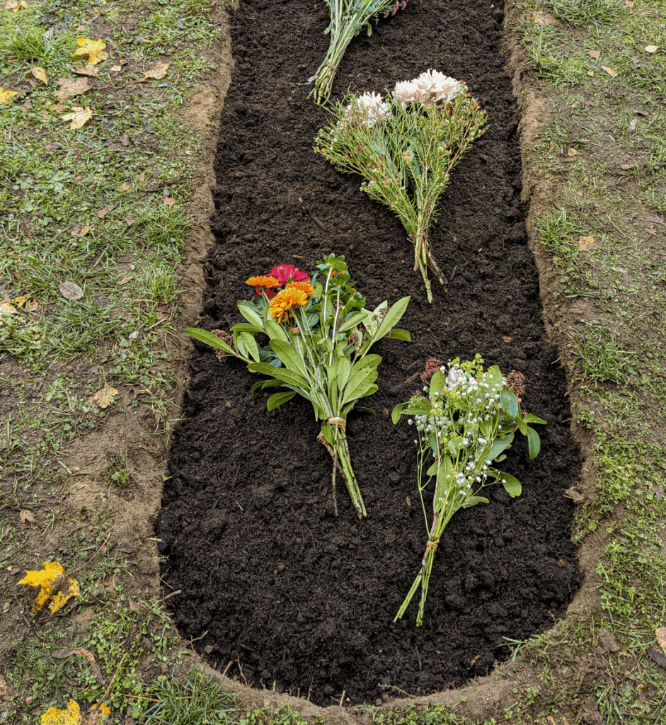 Auf dem Foto ist ein frisch aufgeschüttetes Grab zu sehen, das mit dunkler, feuchter Erde bedeckt ist. Auf der Erde liegen mehrere Blumensträuße, die liebevoll niedergelegt wurden – sie bestehen aus unterschiedlichen, bunten Blüten. Das Grab ist rechteckig, umgeben von kurzgeschnittenem Gras, und am Rand liegen einige herbstliche, gelbe Blätter. In der rechten unteren Ecke steht der Schriftzug „MEINE ERDE“.