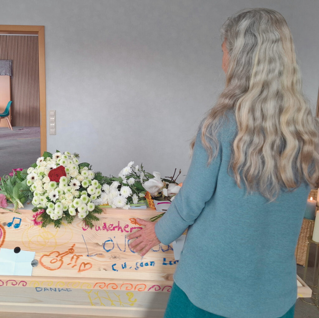 Frau mit langen graues Haar steht beim bunt angemalten geschlossenen Sarg
