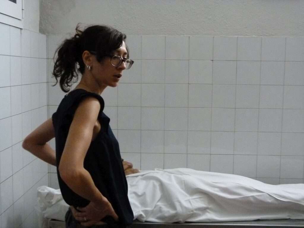 Helena steht vor einem in ein weißes Tuch gehüllten Toten in einem weiß gekachelten Raum