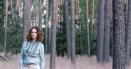 Helena Giuffrida, Gründerin von Atelier Magnolia Bestattungen Berlin, steht vor einem Wald. Sie trägt ein hellblaues Leinenhemd und rosafarbene Hosen. Das Bild wirkt wie ein Gemälde.