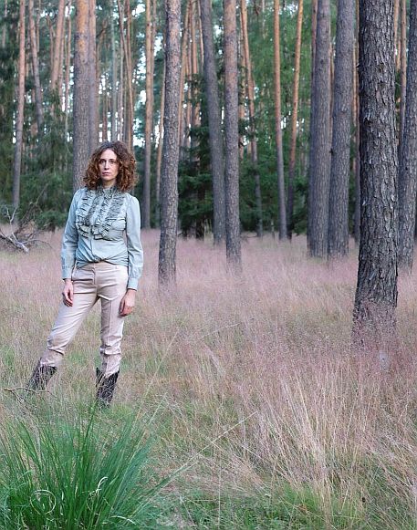 Helena steht in hohem Gras zwischen Bäumen in einem Wald und blickt in die Ferne. Sie trägt eine hellblaue Rüschenbluse, eine helle Hose und Cowboystiefel.