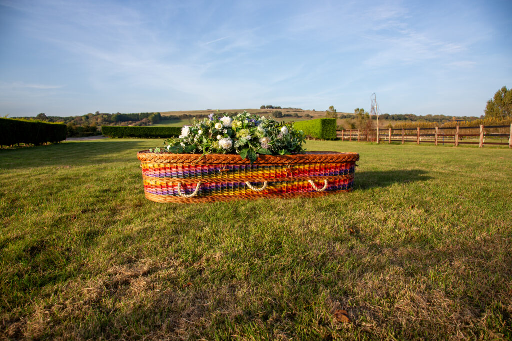 Ein Sarg aus geflochtenen Weide von atlier MAgnolia Bestattungen liegt auf einer Wiese und ist Regenbogenfarbig angemalt