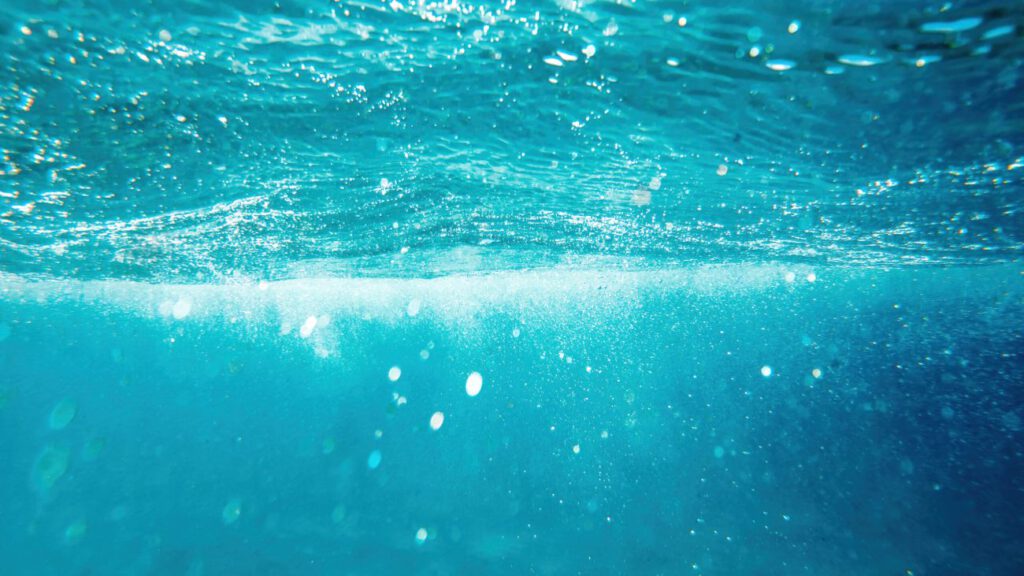 Unterwasseraufnahme, blaues Wasser kurz unter der Oberfläche, die Sonne scheint durch und oben ist eine Wellenstruktur zu erkennen.
